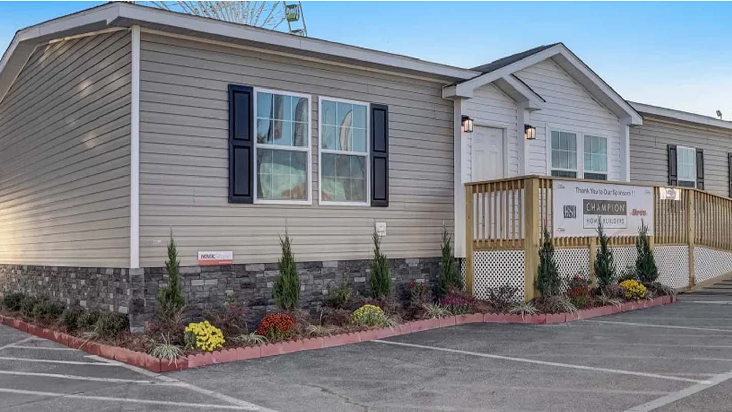 A beige modular home with stone trim, a front porch, and landscaped flowerbeds by a parking lot.